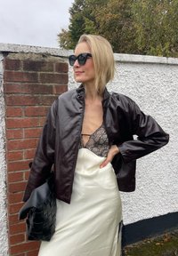 Casaco de pele sintética castanho com zip, sobreposto a uma blusa de renda preta e uma saia de cetim branca. Bolsa tecida preta como acessório.