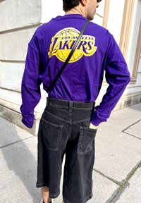 T-shirt a maniche lunghe viola con logo giallo "Los Angeles Lakers" sul retro, abbinato a pantaloni corti in denim neri con due tasche posteriori.