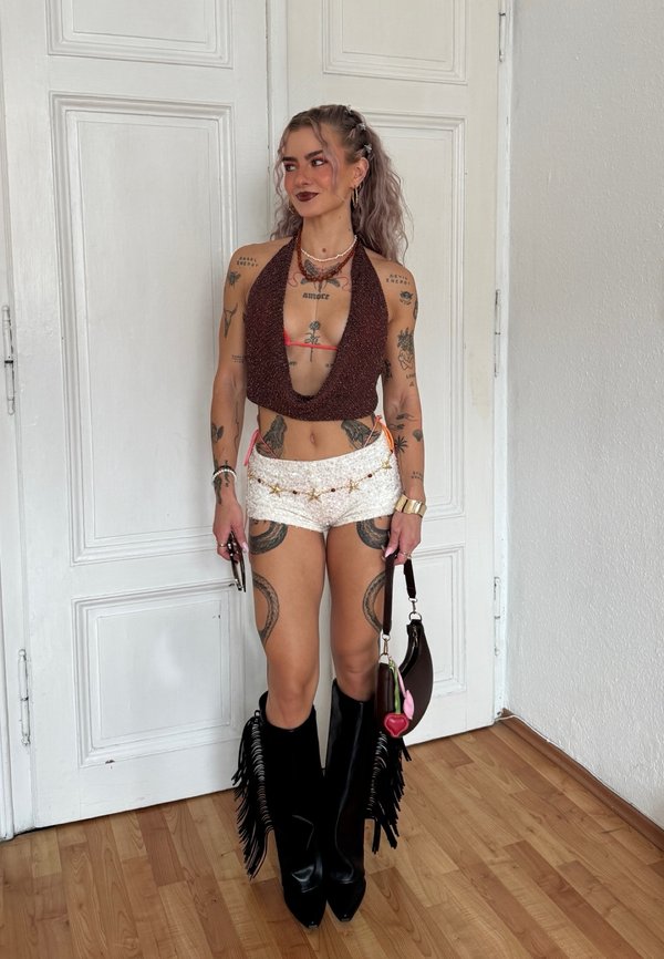 Vrouw met lang krullend haar draagt een haltertop, witte shorts met een sterrenriem, hoge zwarte laarzen met franjes en houdt een kleine bruine handtas binnen.