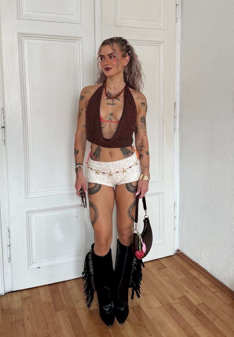 Femme aux longs cheveux bouclés porte un haut dos-nu, un short blanc avec une ceinture étoilée, de grandes bottes noires à franges et tient un petit sac à main marron à l'intérieur.