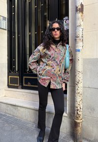 Femme aux cheveux bouclés et foncés portant des lunettes de soleil noires, une chemise à motif cachemire, un pantalon noir, debout près d'une porte noire et d'un poteau rouillé dans la rue.