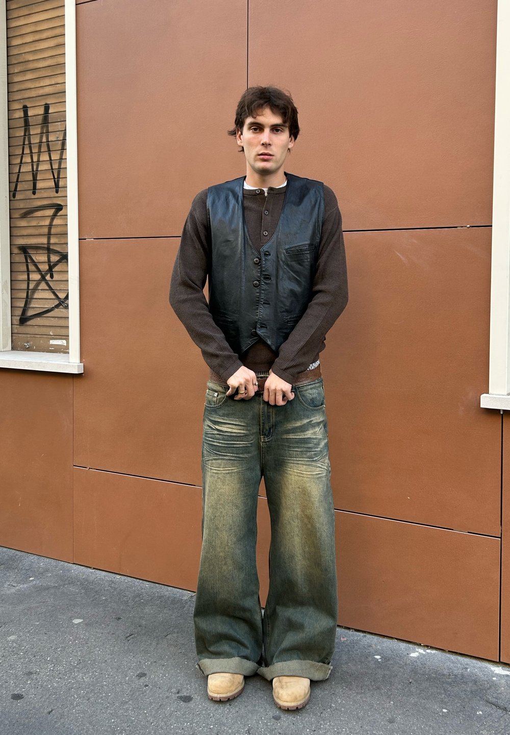 Chaleco de cuero negro sobre una camisa de manga larga marrón, combinado con vaqueros de mezclilla anchos azules y zuecos color tostado, en un fondo de pared marrón.