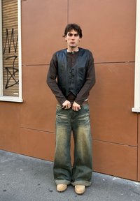 Chaleco de cuero negro sobre una camisa de manga larga marrón, combinado con vaqueros de mezclilla anchos azules y zuecos color tostado, en un fondo de pared marrón.