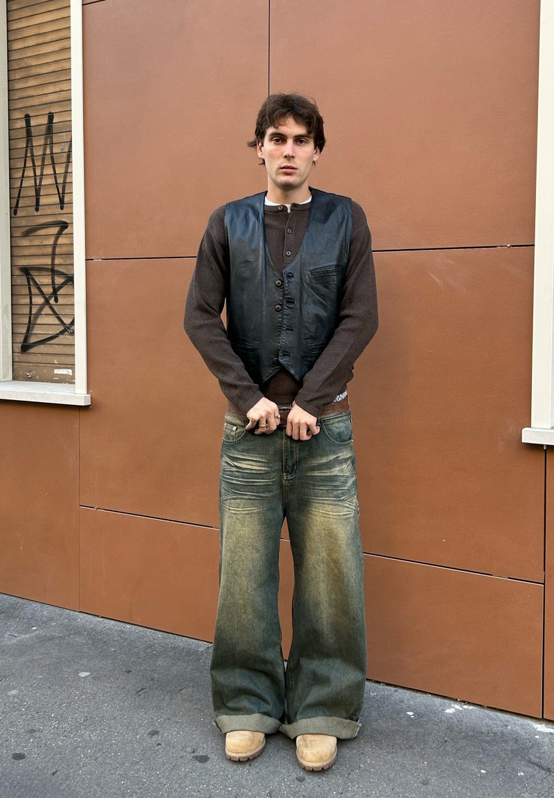 Schwarze Lederweste über einem braunen Langarmshirt, kombiniert mit weiten blauen Jeans und beigen Clogs, vor einer braunen Wand.