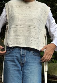 Top sem mangas, tricotado na cor creme, com padrões texturizados, motivos de corações e laços nas laterais, usado sobre uma camisa de manga longa branca.