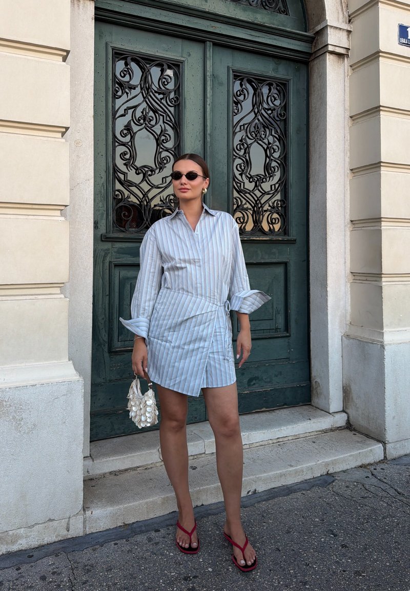Femme en robe chemise rayée, lunettes de soleil et sandales rouges, debout tenant un petit sac à main blanc devant des portes doubles décoratives vertes.