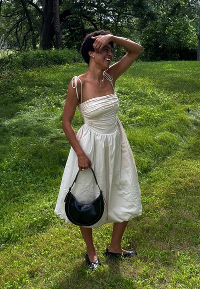 Vestido color crema con cuerpo fruncido, tirantes finos y falda amplia. Bolsa negra con asa curvada. Fondo de césped.