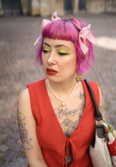 Vestido corto rojo con un acento de lazo. Pelo rosa con cintas, sombra de ojos verde intensa y labios rojos. Múltiples tatuajes visibles en el brazo y el pecho.