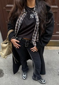 Sort stor frakke, ternet skjorte over en sort crop top, sorte jeans med hvide syninger og sorte/sølv sneakers. Beige taske og synlige ringe.