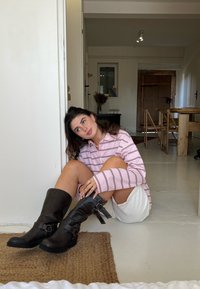 Botas de couro pretas até ao joelho com fivelas e tiras, combinadas com uma camisa rosa às riscas e calções brancos, sobre um tapete texturizado num espaço iluminado.
