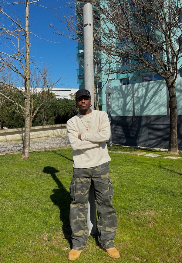 Uomo con felpa beige, pantaloni mimetici e cappello nero, in piedi con le braccia incrociate appoggiato a un palo in un parco urbano verde in una giornata di sole.