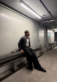 Szürke kardigán fehér trikó fölött, fekete bő szárú nadrág és fekete cipők. Fém korlátnak támaszkodik egy gyengén megvilágított folyosón.