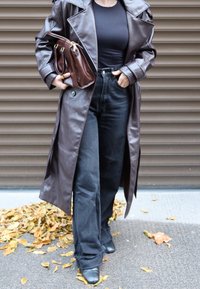 Langer brauner Ledermantel über einem schwarzen, eng anliegenden Oberteil, kombiniert mit hoch taillierten schwarzen Jeans mit weitem Bein und schwarzen Stiefeln mit Absatz. Braune Handtasche.