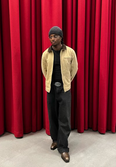 Figura alta vestida con una chaqueta beige de cuello con detalles marrones, camiseta negra, jeans oscuros de pierna ancha y zapatos marrones, de pie frente a unas cortinas rojas.