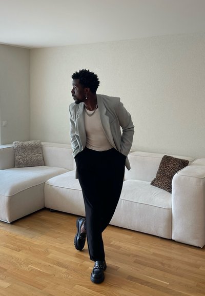 Hombre con pendientes, collar, blazer claro y pantalones negros, de pie con las manos en los bolsillos en una sala de estar minimalista con sofá blanco y suelo de madera.
