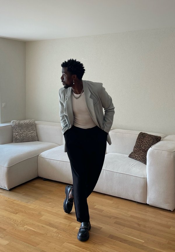 Man with earrings, necklace, light blazer, and black pants standing with hands in pockets in a minimalist living room with white sofa and wooden floor.