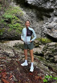 Svijetloplavi i crni zip-jakna s geometrijskim uzorkom, maslinasto zeleni šorts i svijetloplave sportske cipele na stjenovitom, prirodnom terenu.