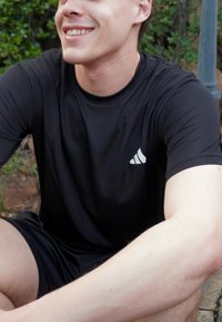 Jeune homme portant un t-shirt et un short de sport Adidas noirs, assis en plein air, souriant, avec un arrière-plan de verdure.