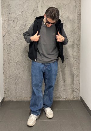 Chaleco negro con cremallera sobre una sudadera gris, combinado con unos vaqueros de corte amplio en azul claro y zapatillas en tonos neutros. Joyas plateadas llamativas como acento.