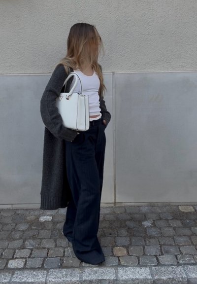 Mujer de pie en una calle empedrada, vestida con una camiseta sin mangas blanca, pantalones anchos oscuros, un cárdigan largo oscuro y sosteniendo un bolso blanco.