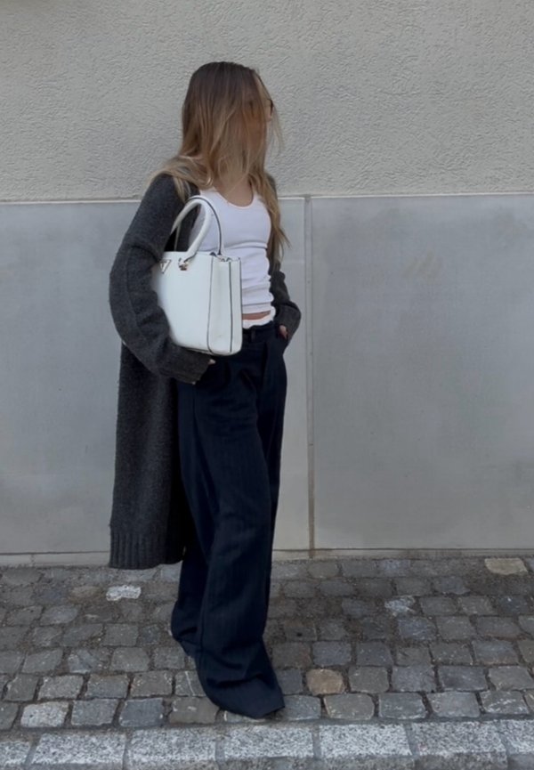 Kobieta stoi na brukowanej ulicy, ubrana w biały top na ramiączkach, ciemne szerokie spodnie, długi ciemny sweter i trzyma białą torebkę.