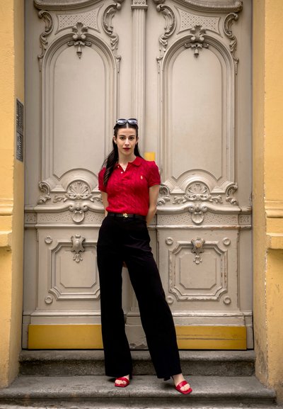 Camisa de polo de punto rojo, pantalones negros de talle alto y sandalias de plataforma rojas. De pie frente a una puerta ornamentada de color claro.
