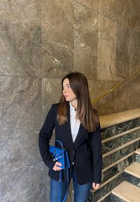 Blazer nero sopra una camicia bianca, che tiene una piccola borsa blu. Lunghi capelli castani, in piedi vicino a un muro di marmo e a una scalinata.