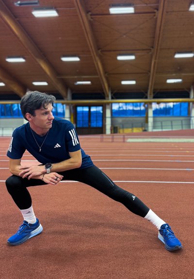 Atleta con camiseta Adidas azul marino con rayas blancas, leggings negros y zapatillas azules, estirándose en una pista interior roja con techo de madera.
