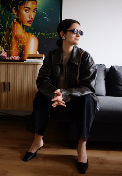 Chaqueta de cuero negra, blusa beige y pantalones anchos negros. Tacones de punta negra. Gafas de sol oscuras. Fondo moderno con arte.