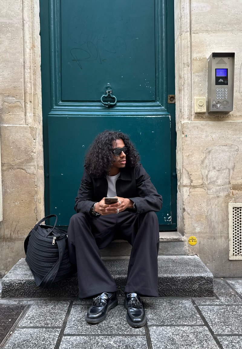 Personne aux cheveux bouclés et lunettes de soleil assise sur une marche en pierre tenant un téléphone, à côté d'un sac noir, devant une grande porte verte avec un interphone à proximité.