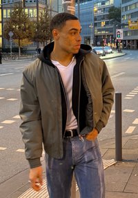 Jeune homme aux cheveux courts et taches de rousseur portant une veste verte, une chemise blanche et un jean, debout sur un trottoir en ville près d'un poteau de rue.