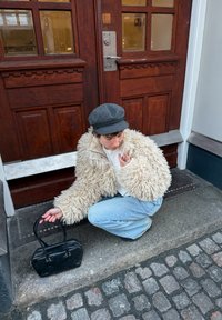 Personne accroupie près de portes doubles en bois, portant une veste crème en fourrure, une casquette grise, un jean bleu clair, tenant un sac à main noir sur une marche en pierre.