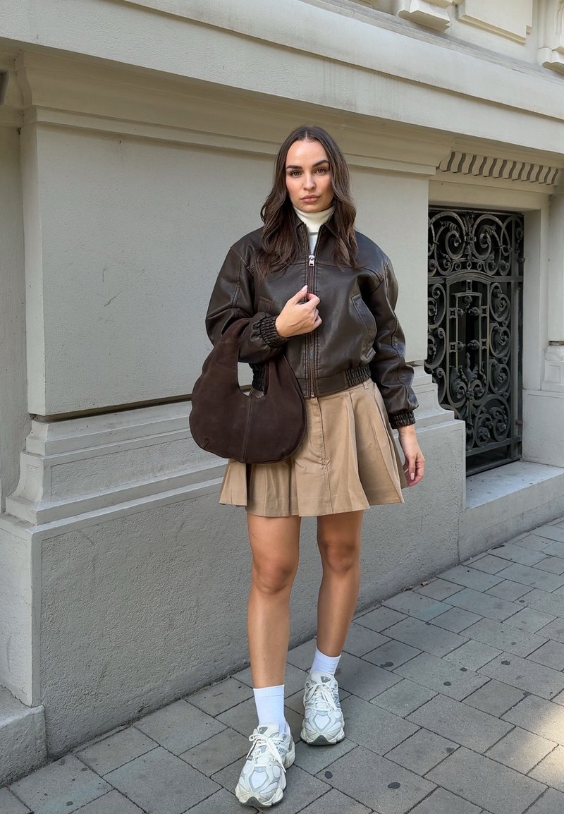 Femme portant une veste en cuir marron foncé, une jupe plissée beige, des baskets blanches et tenant un sac à main marron foncé, debout sur un trottoir urbain.