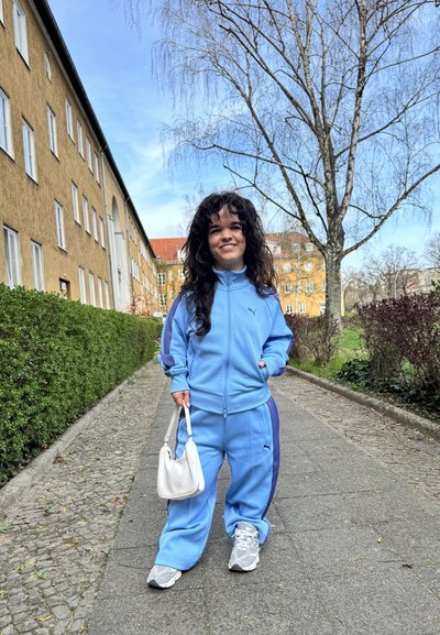 Chándal de color azul claro con franjas laterales moradas, compuesto por una chaqueta con cremallera y pantalones a juego. Bolso blanco y zapatillas grises. Entorno al aire libre.