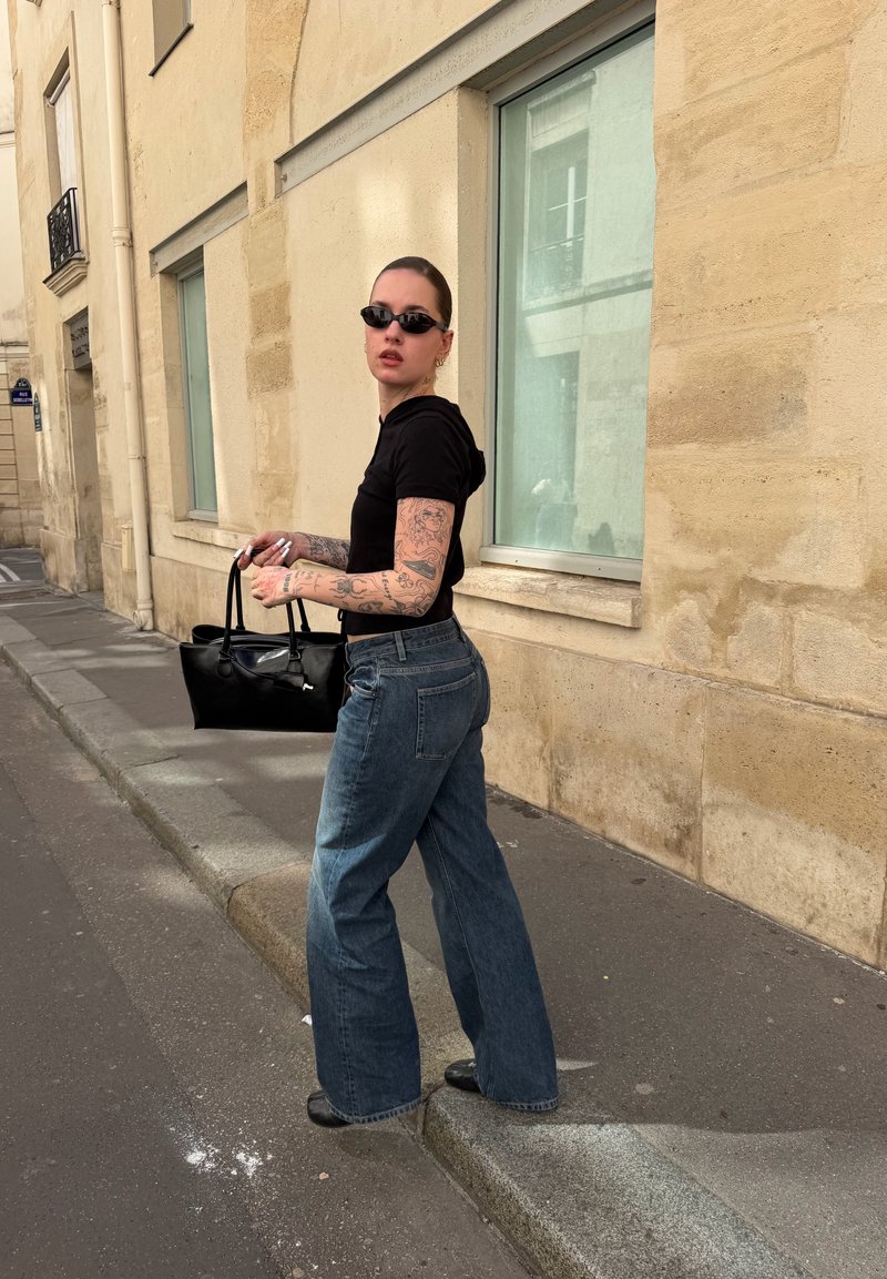 Femme avec des tatouages portant un haut noir et un jean large, tenant un sac à main noir, debout sur un trottoir en ville près d’un bâtiment en pierre beige.