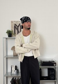 Bēša kapuces džemperis ar rāvējslēdzēju, uzvilkts virs baltas t-krekla, melnas bikses, melna bandana, redzamas tetovējumi. Aizmugurē esošajā plauktā izvietoti augi un ieraksti.
