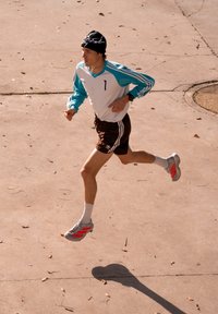 Jeune homme courant sur une surface en béton fissurée, portant un t-shirt à manches longues bleu sarcelle et blanc, un short noir, des baskets grises et un bonnet noir.