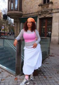 Femme aux cheveux bouclés portant une casquette orange, un haut rose sur un sous-pull gris, une jupe blanche à fines rayures et des baskets, appuyée sur une rambarde en verre en plein air.