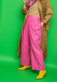 Personne portant un pantalon large rose, des bottines jaunes à bout pointu, un haut beige, un manteau marron et une écharpe à motifs, se tenant devant un fond vert.