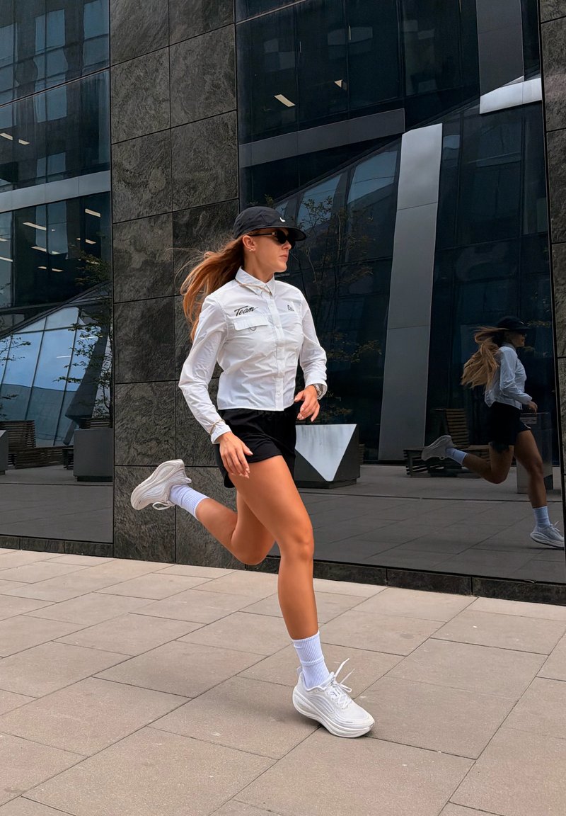 Femme portant une veste blanche, un short noir, une casquette et des lunettes de soleil, faisant du jogging sur une surface extérieure pavée près d'un bâtiment en verre réfléchissant.