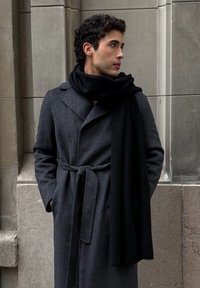 Jeune homme aux cheveux bouclés, debout contre un mur de pierre, portant un manteau gris foncé ceinturé et une longue écharpe noire, regardant sur le côté.