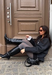 Cizme negre din piele până la genunchi, dresuri gri, palton negru supradimensionat și o geantă mică neagră. Ușă texturată maro în fundal.