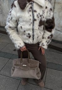 Person iført en hvid og brun plettet pelsjakke, brune bukser og støvler, der holder en stor taupe ruskinds håndtaske på en byvej.