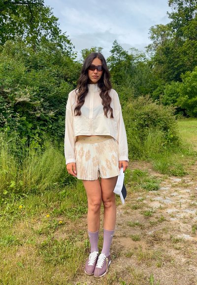 Mujer con cabello largo que lleva gafas de sol, chaqueta ligera, pantalones cortos estampados, calcetines morados y zapatillas, de pie en un sendero con vegetación de fondo.