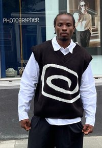 Gilet lavorato a maglia maxi di color marrone scuro, con un motivo a spirale bianca, indossato su una camicia bianca con colletto, abbinato a pantaloni neri larghi.