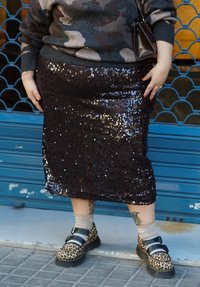 Personne portant une jupe sombre à sequins, un pull à motif camouflage, des chaussures léopard à brides, des chaussettes beiges et des tatouages visibles sur les jambes.