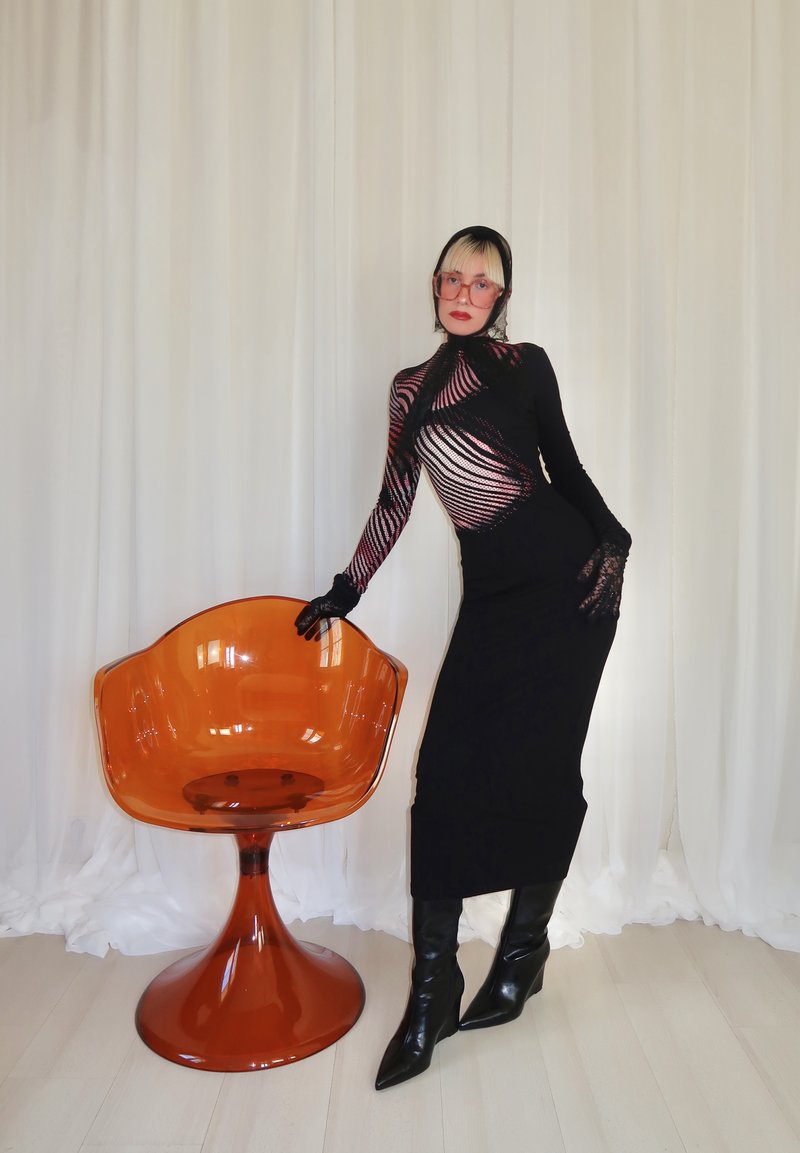 Person in black high-neck dress with sheer striped sleeves and gloves, posing beside an orange modern swivel chair on a light floor.