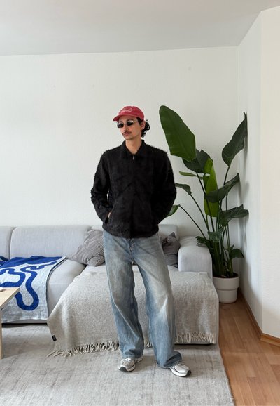 Hombre con gorra roja, chaqueta negra mullida, gafas de sol, vaqueros anchos y zapatillas de deporte está de pie en un salón con sofá gris y planta alta verde.