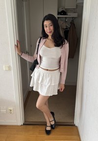 Weißer gestufter Rock mit Stickerei, weißes Tanktop, pinkfarbener Cardigan mit Knöpfen und schwarze Mary Jane-Schuhe. Steht drinnen in der Nähe einer Tür.