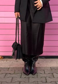 Trench nero sopra una gonna in raso nero e stivali a punta. Borsa nera con tracolla a catena e dettagli decorativi. Sfondo rosa.